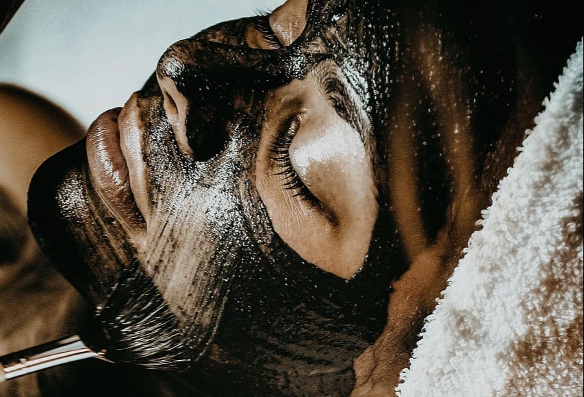 A person lying down while a dark charcoal or clay facial mask is applied to their face with a brush